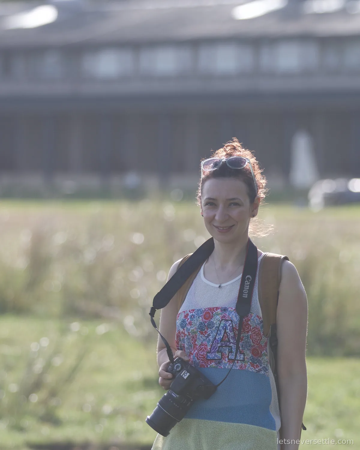 Inga smiling with sunglasses pushed up into her hair and a Canon DSLR on a strap around her neck, green grassland blurred behind