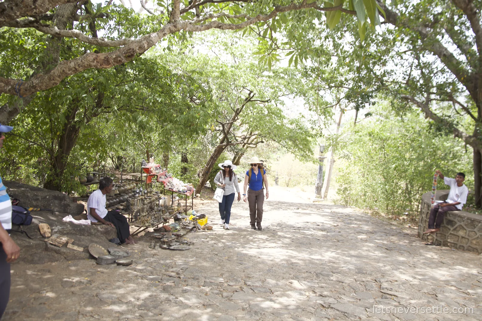 A shaded stone path at Dambulla lined with a stall of brass lamps, plates and carved masks, Inga and a friend walking up in sun hats