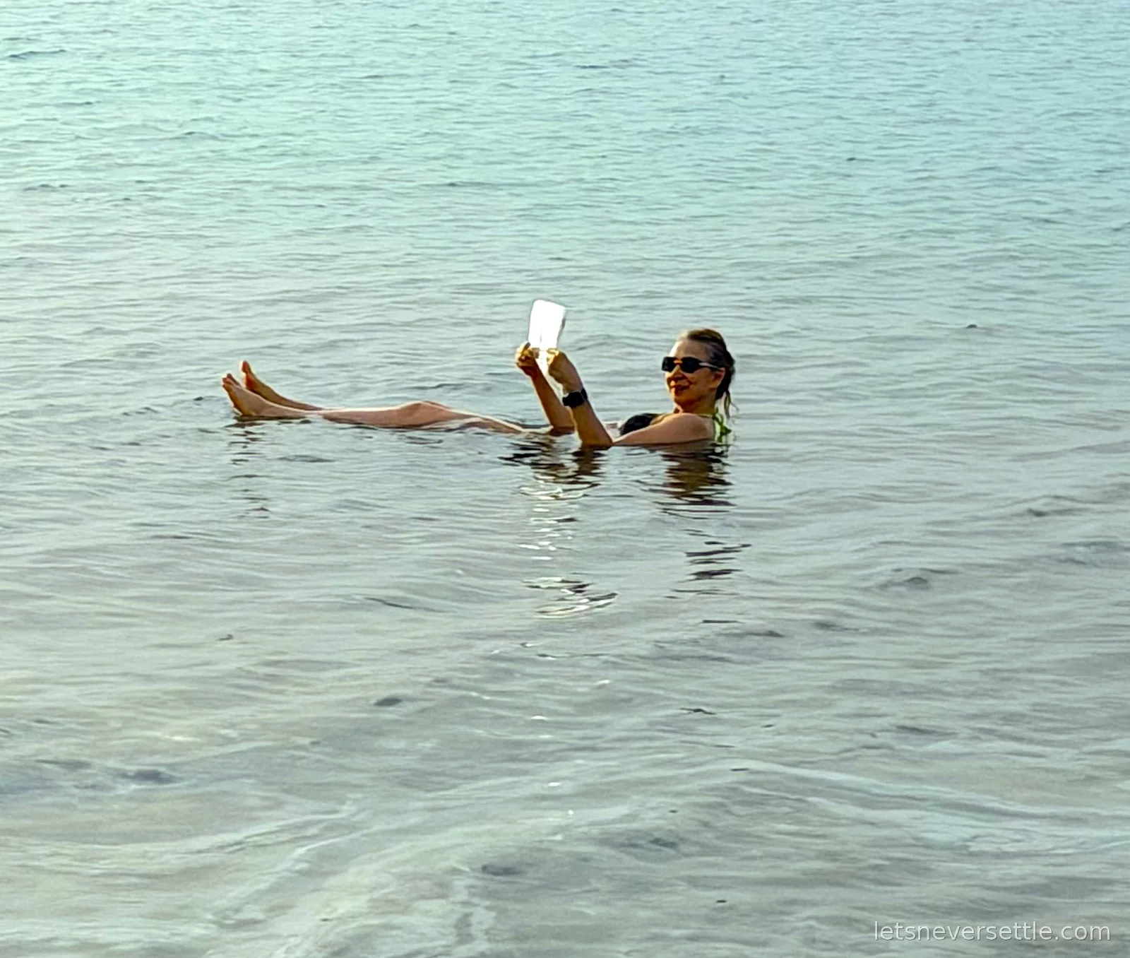 Inga floating on her back in the pale green Dead Sea with her feet up, holding a white card above her face to read