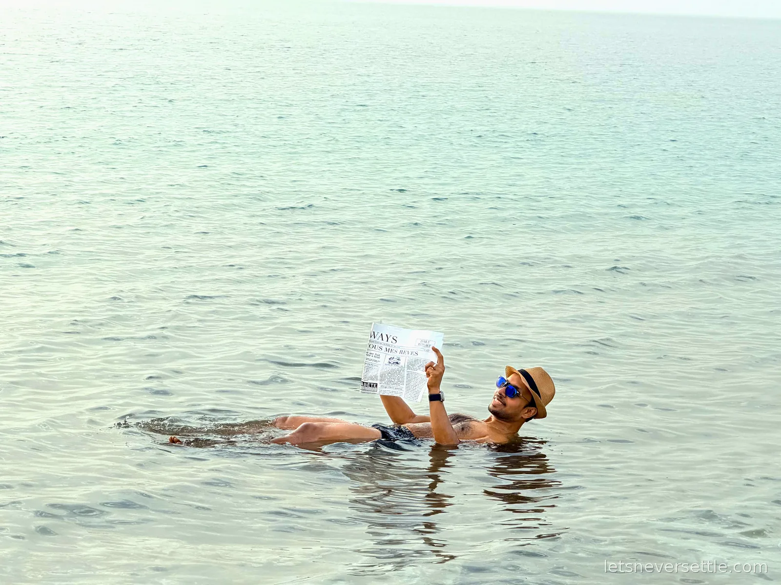 Dileep in a straw hat and sunglasses floating on his back in the pale green Dead Sea, reading a newspaper