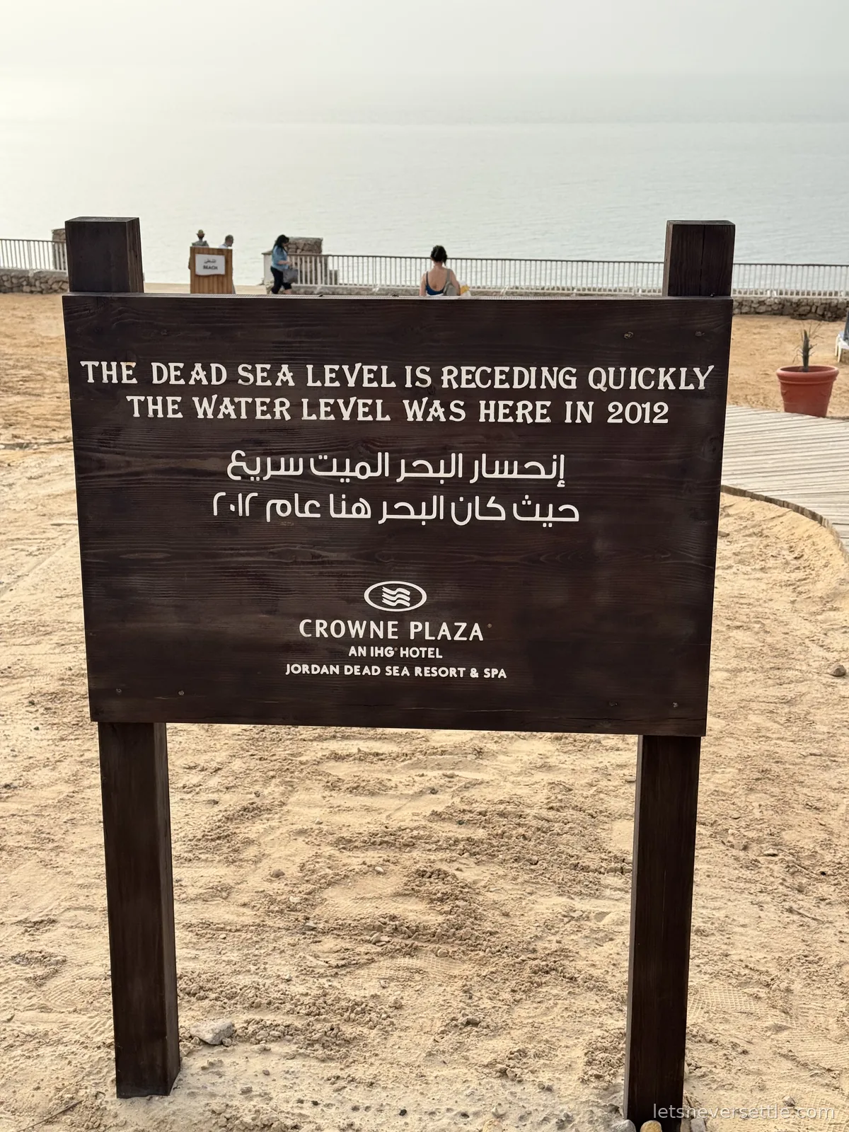 A wooden sign on the beach reading The Dead Sea level is receding quickly, the water level was here in 2012, in English and Arabic, with the shoreline far behind it