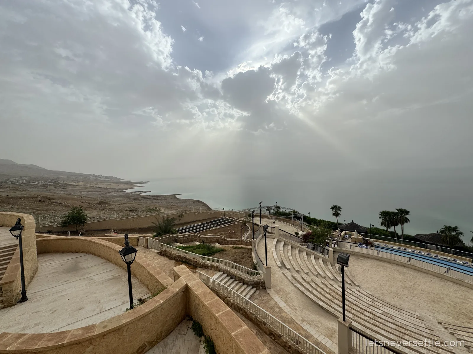 Terraced stone steps and an open-air amphitheatre at a Dead Sea resort, with sunbeams breaking through cloud over the water and the far shore