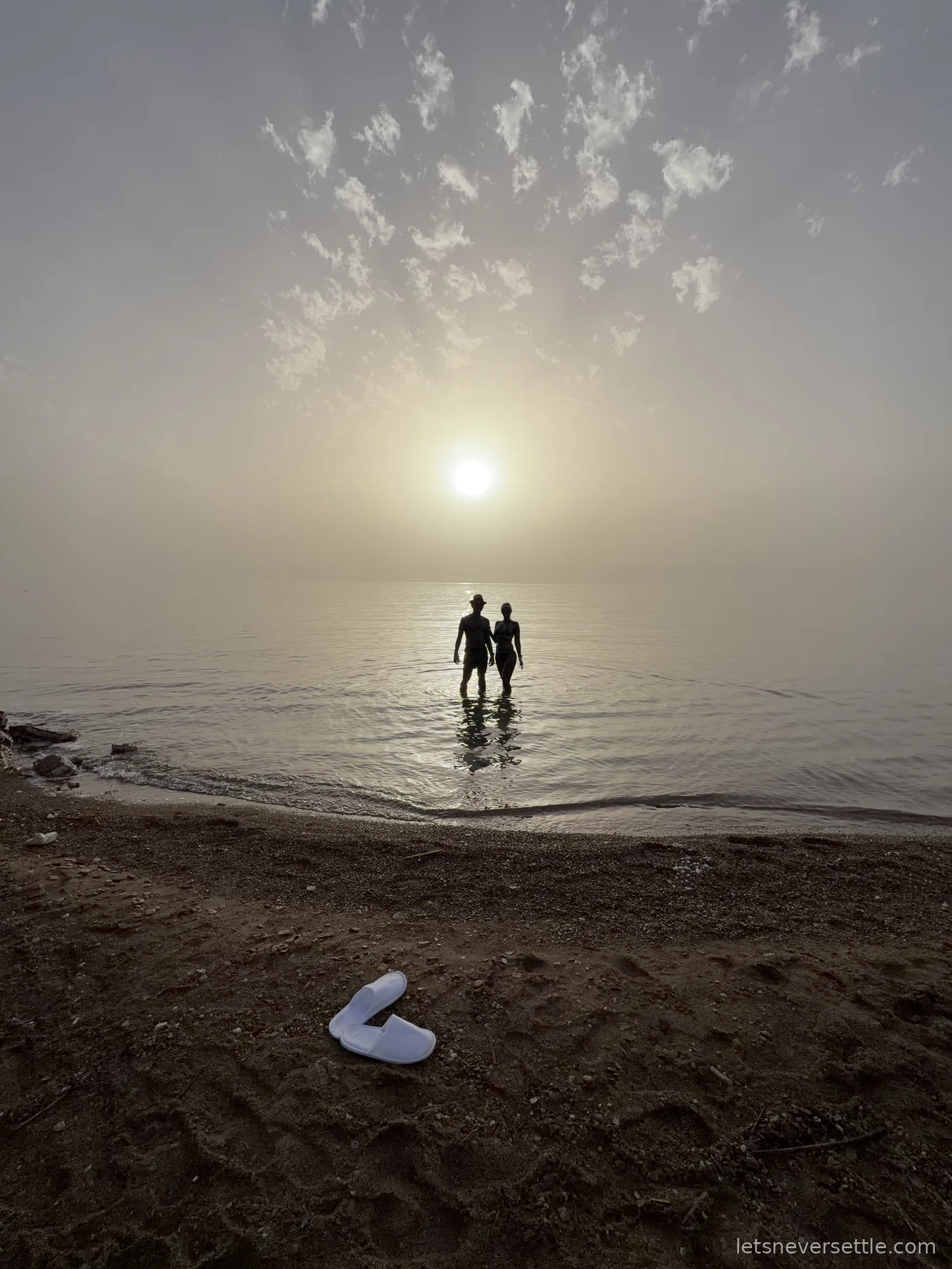 Inga and Dileep silhouetted in the shallows of the Dead Sea at sunset, with a pair of white slippers left on the dark pebble beach
