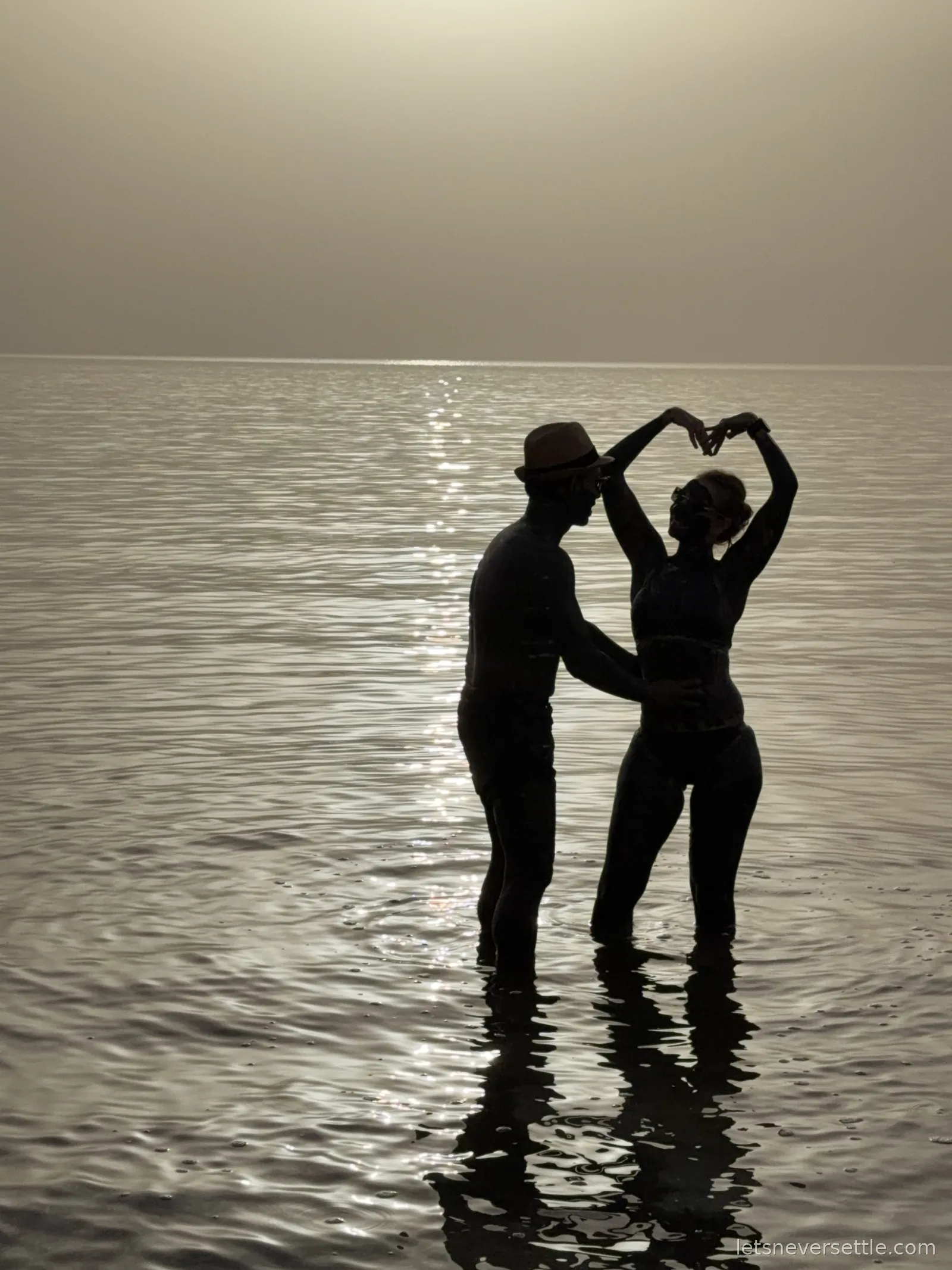 Silhouettes of Inga and Dileep standing in the glittering Dead Sea at sunset, raising their hands together above their heads