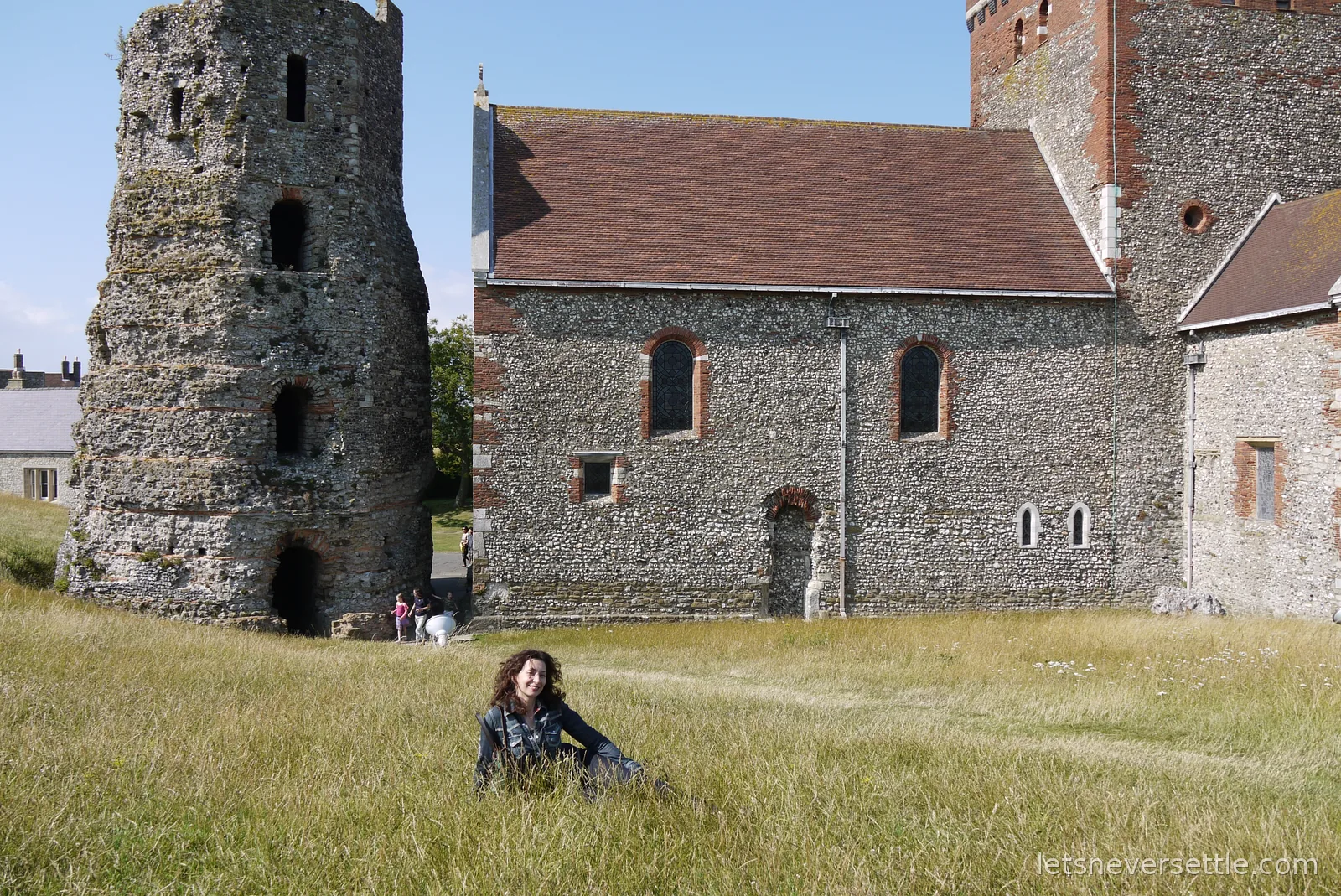Inga sitting in long golden grass at Dover Castle in front of the ruined Roman pharos lighthouse and the flint church of St Mary-in-Castro