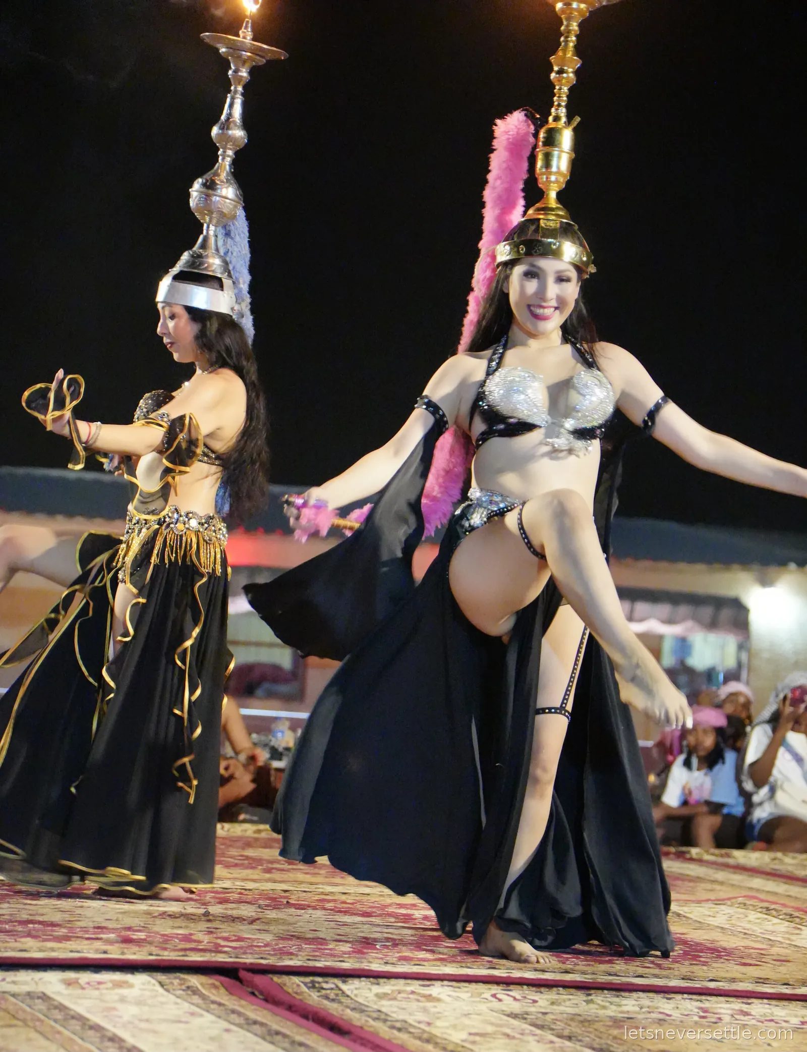 Two dancers in black and gold costumes performing at the desert camp, each balancing an ornate candelabra headpiece