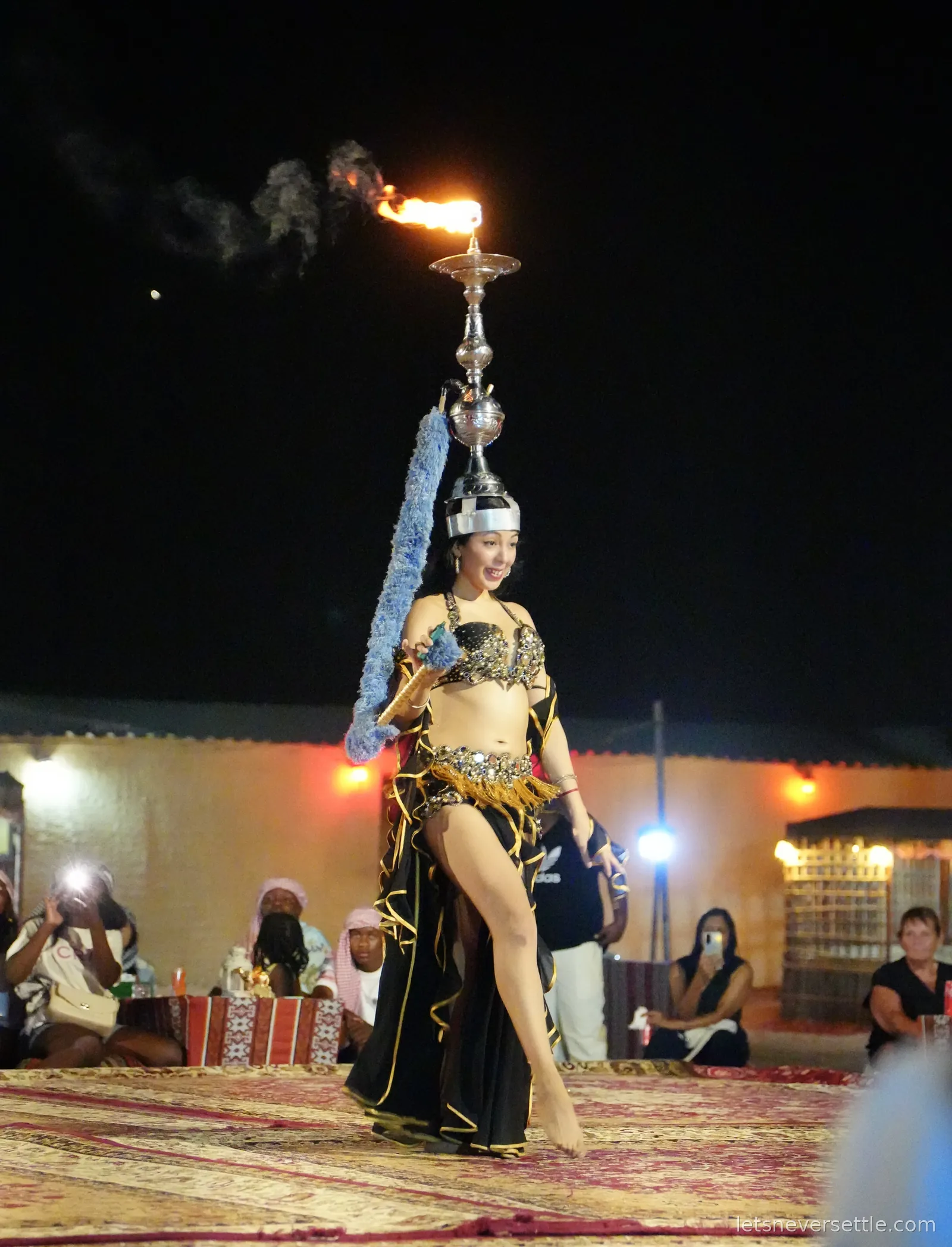 A dancer at the desert camp balancing a tall lit candelabra on her head, the flame burning above her in the dark