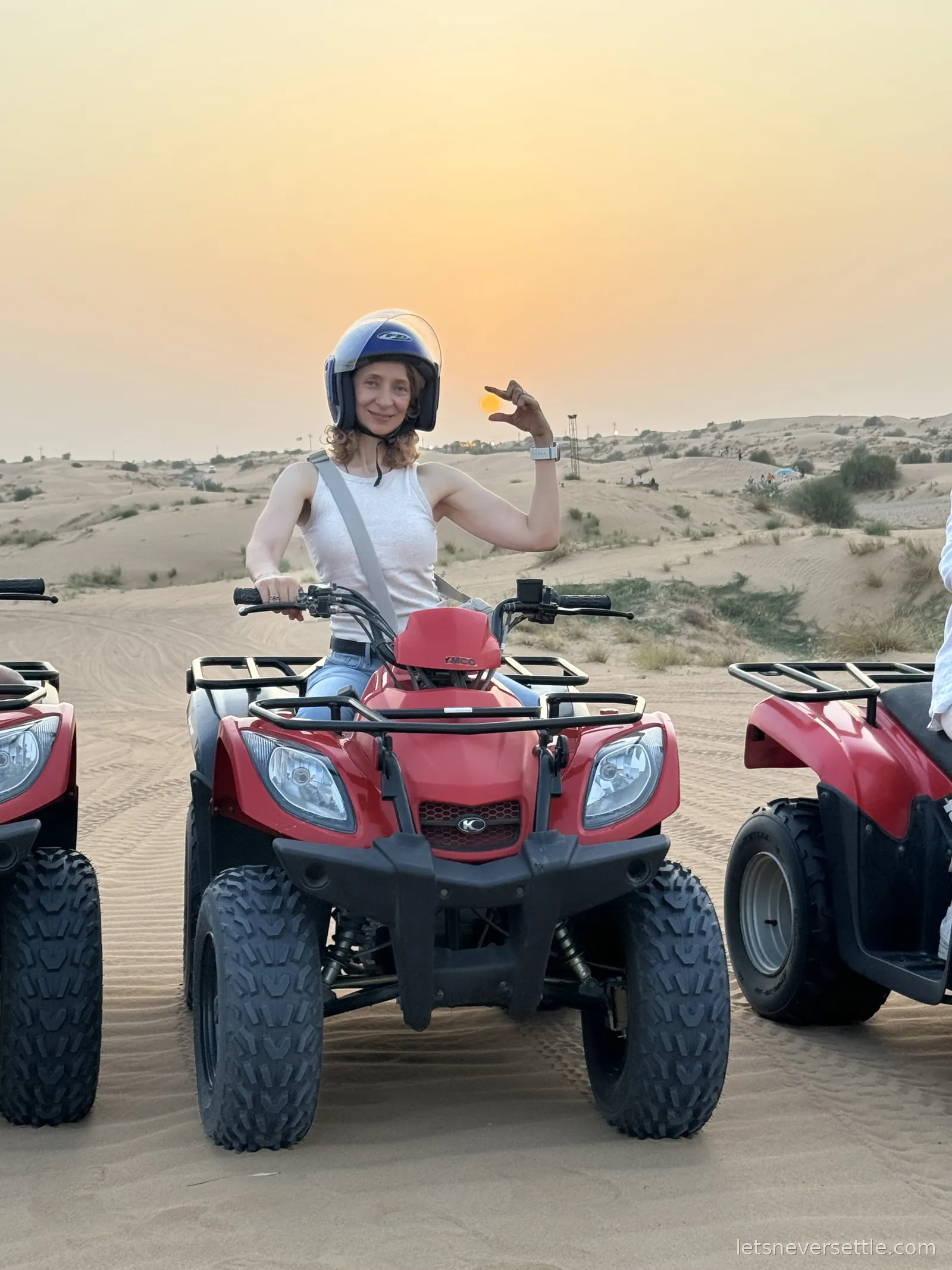 Inga in a helmet sitting on a red quad bike on the sand at sunset, more quad bikes lined up beside her and pale dunes behind