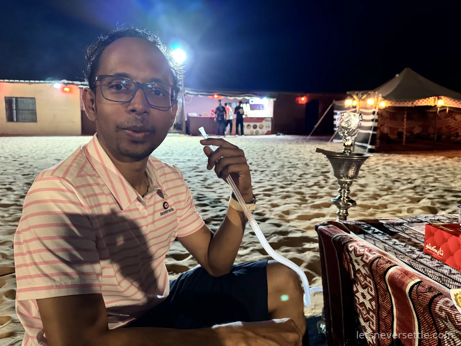 Dileep sitting on a low patterned cushion at the desert camp at night holding a shisha pipe, a lit tent and stage behind him