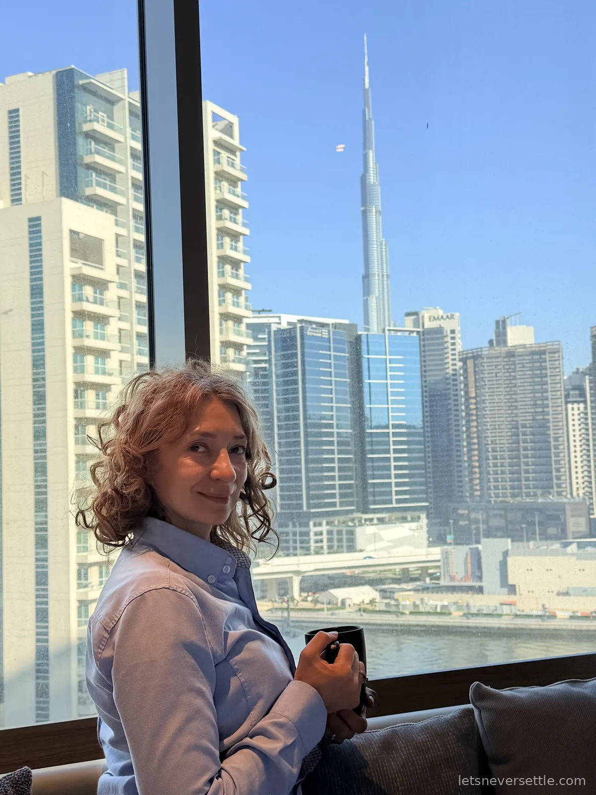 Inga sitting with a coffee mug by a hotel window in daylight, the Burj Khalifa and the towers of Business Bay through the glass