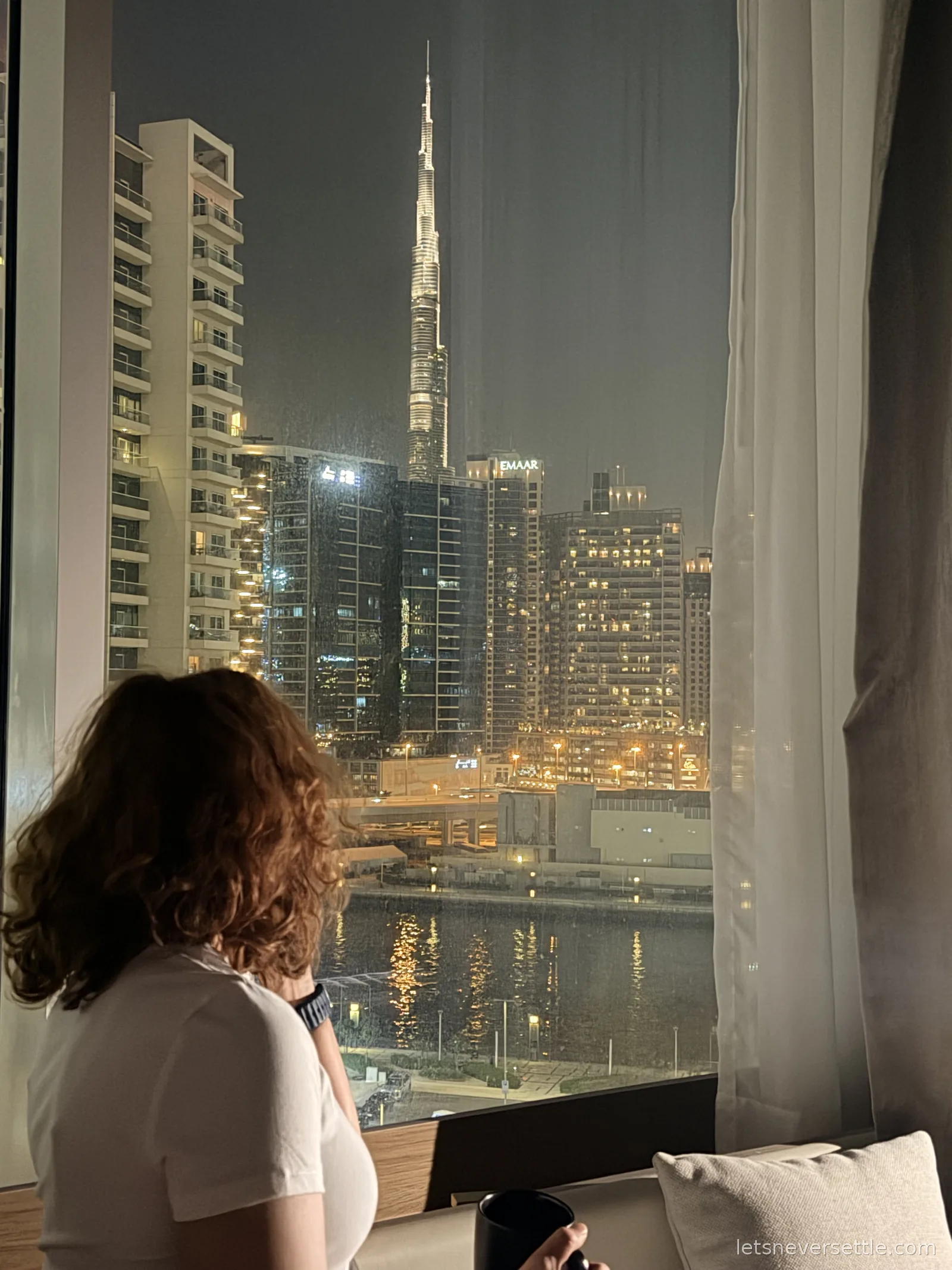 Inga looking out of a hotel window at night towards the illuminated Burj Khalifa and the towers of Downtown Dubai