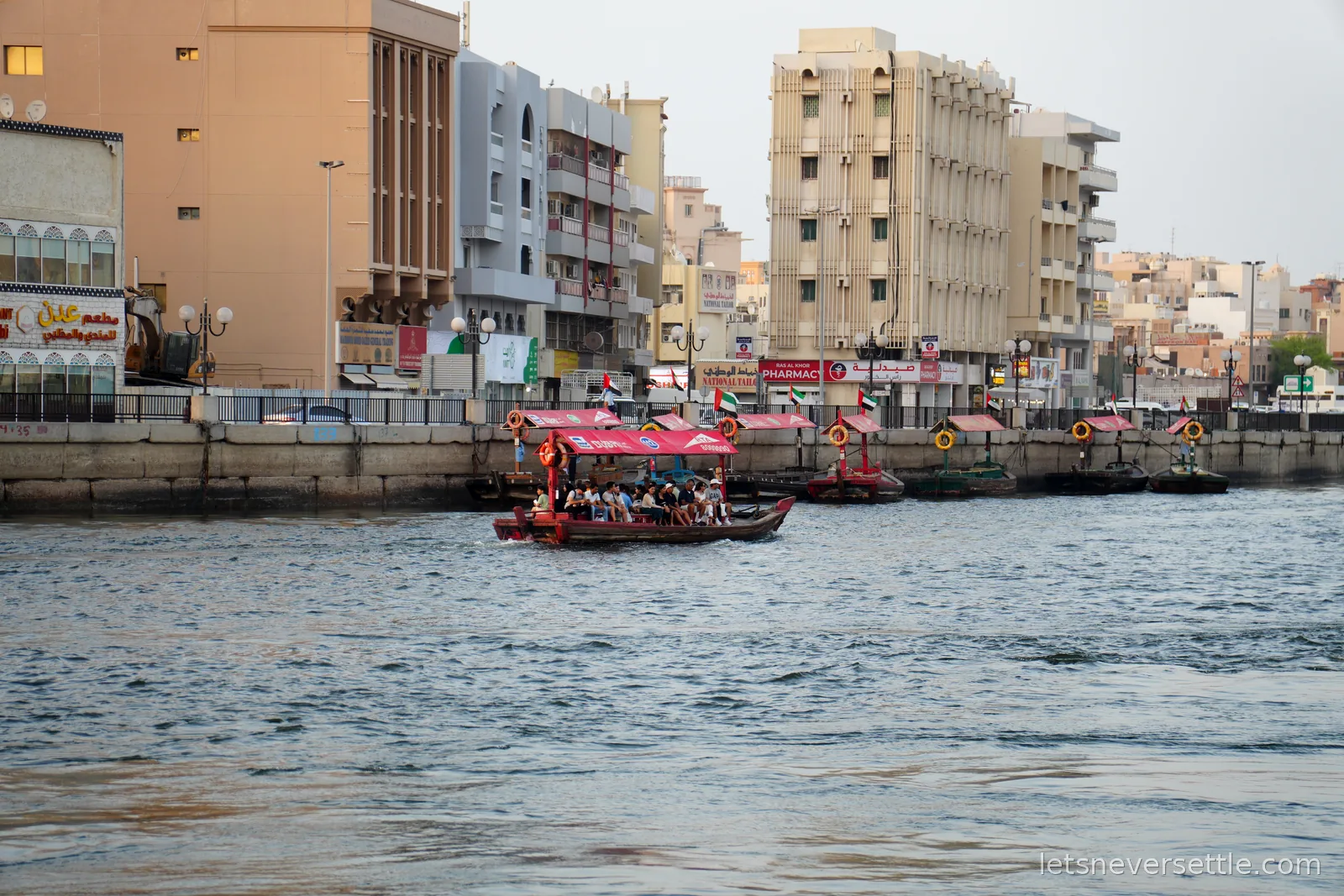 A wooden abra ferry carrying passengers across Dubai Creek, UAE flags flying over the moored boats and old Deira buildings behind