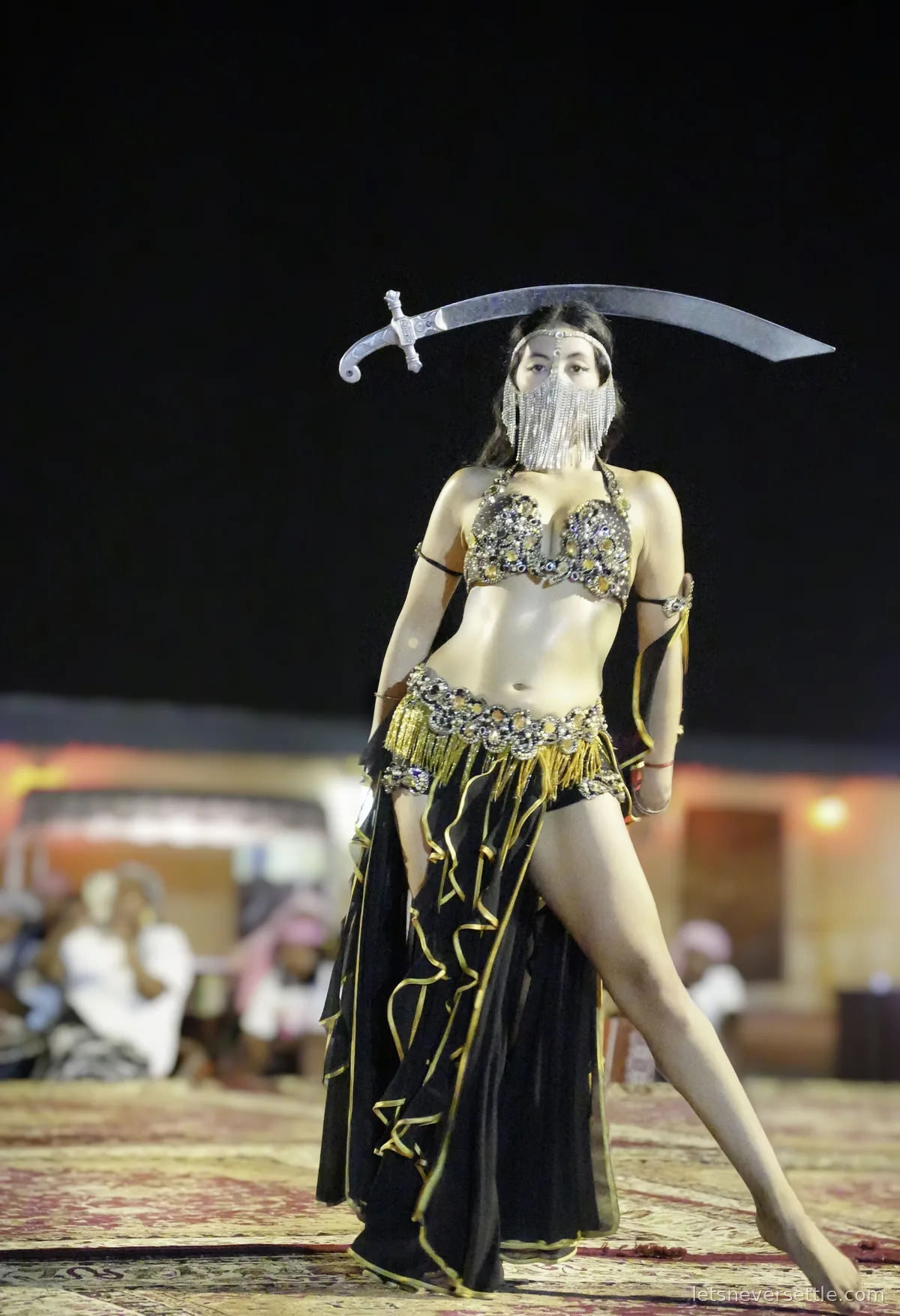 A dancer in a black and gold costume with a face chain veil balancing a curved sword across her head at a desert camp show