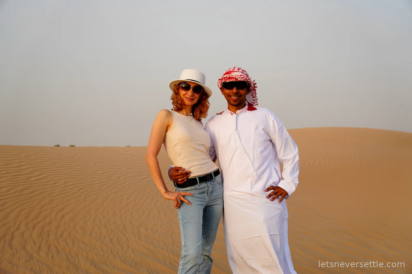 Inga in a white top and sun hat with Dileep wearing a white kandura and red checked ghutra headdress, standing on the dunes at golden hour