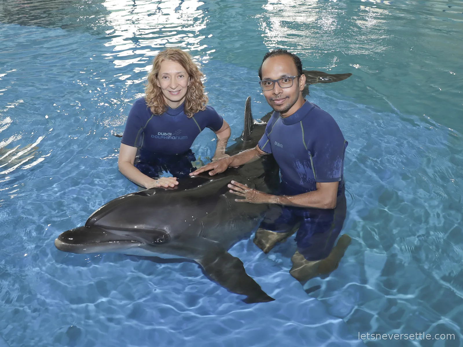 Inga and Dileep in wetsuits kneeling in clear blue water with a dolphin resting across their arms at the Dubai Dolphinarium