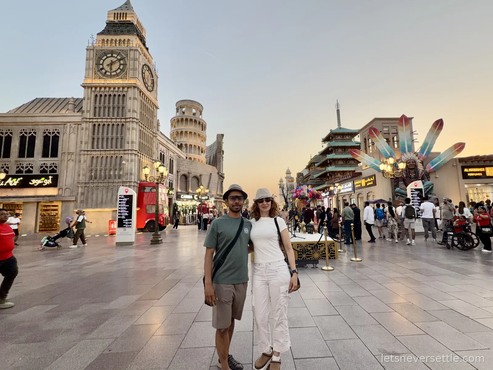 Inga and Dileep standing on the boulevard at Global Village with the replica Big Ben clock tower and leaning tower lit up behind them