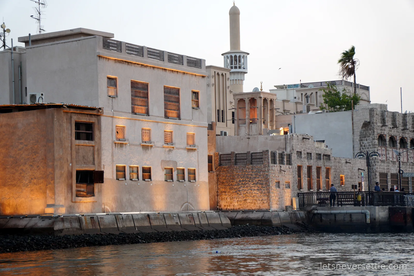 Coral-stone buildings, wind towers and a slim minaret glowing at dusk along the old Shindagha waterfront on Dubai Creek