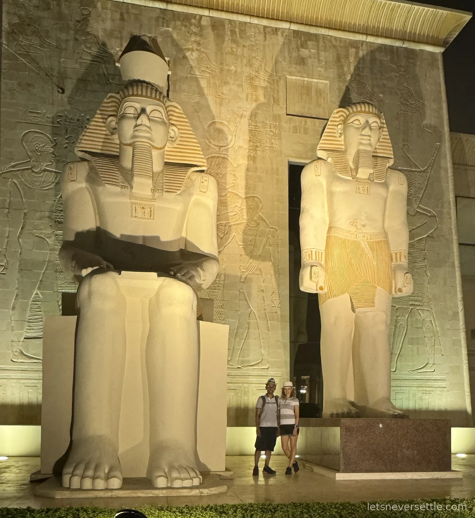 Inga and Dileep standing at night between two giant illuminated pharaoh statues in front of a wall of hieroglyphic reliefs at Wafi, Dubai
