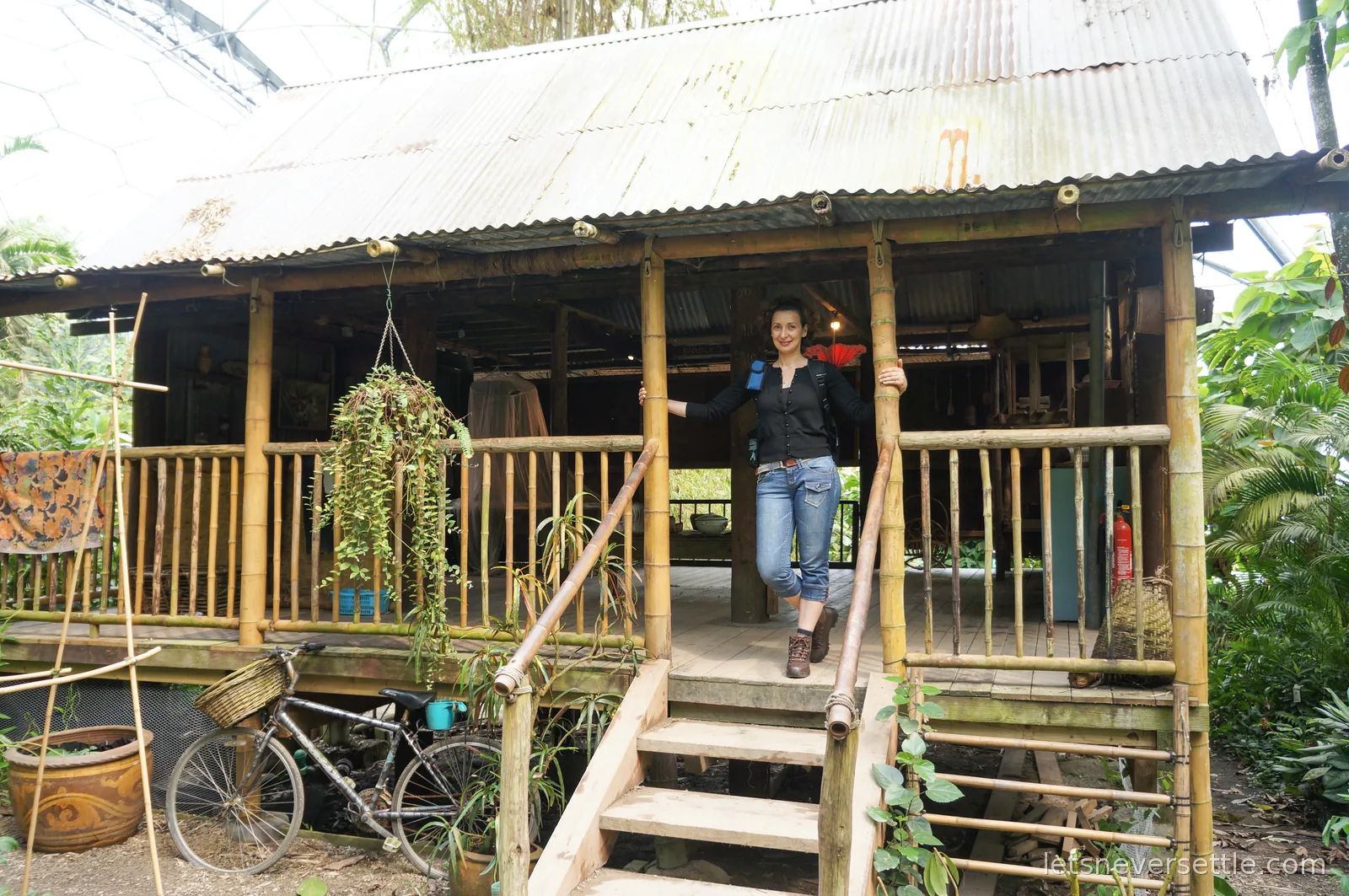 Inga standing on the wooden steps of a bamboo hut with a corrugated roof inside the rainforest biome, hexagonal roof panels visible beyond the treetops