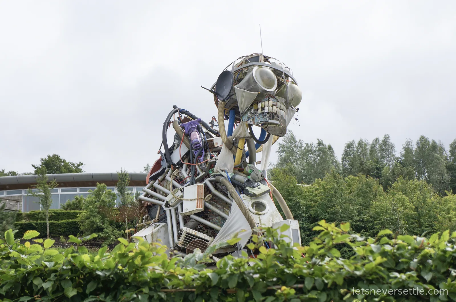 Giant humanoid sculpture assembled from discarded appliances, satellite dishes and machine parts towering over greenery at the Eden Project