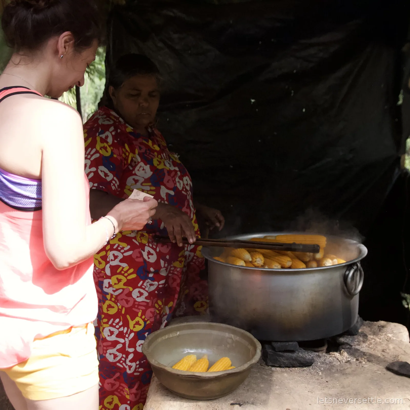 Inga buying corn from a woman ladling cobs out of a huge steaming pot over a wood fire