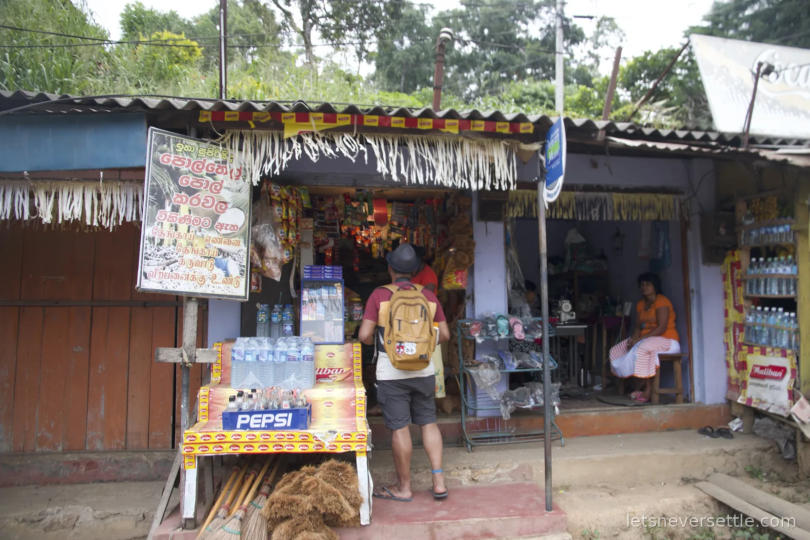 Dileep with a backpack browsing at a small village shop with a Pepsi stand, bottled water and brooms out front