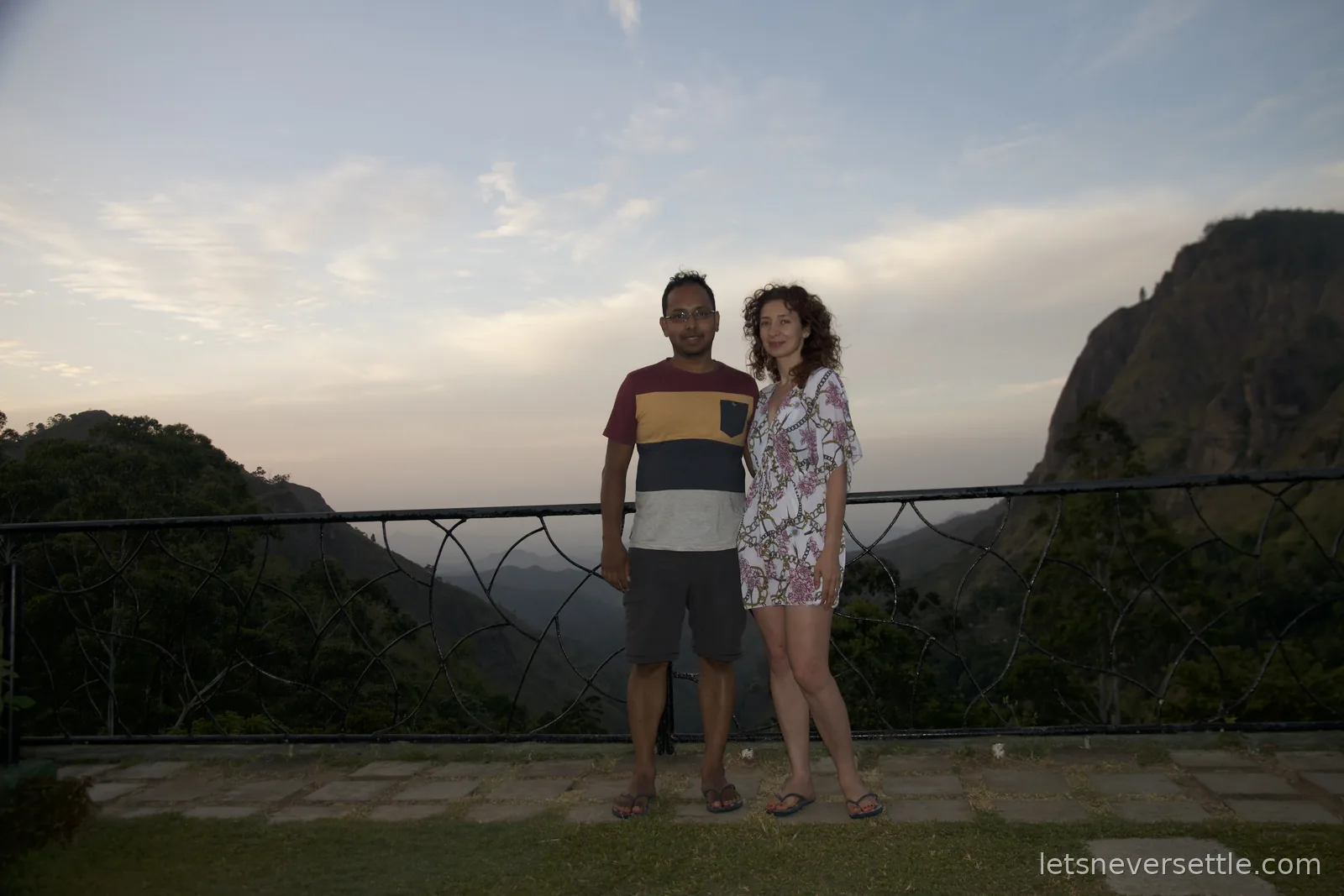 Inga and Dileep standing together at the garden railing at dusk, the ridges of Ella Gap silhouetted behind