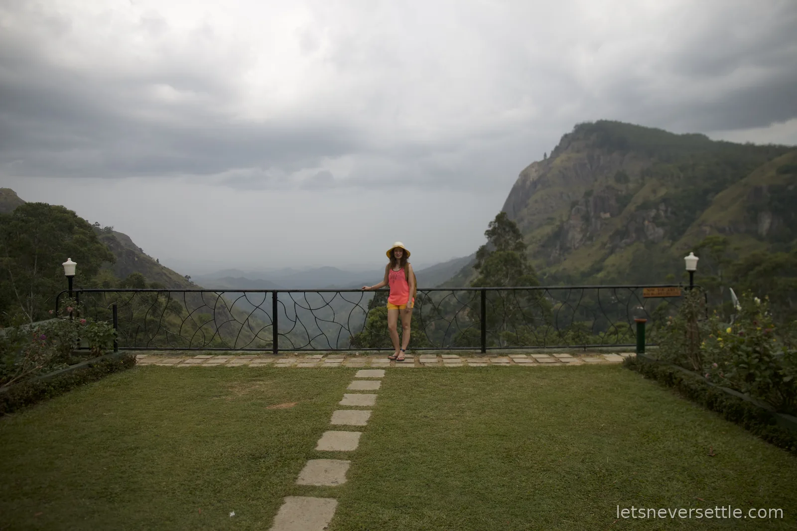 Inga in a sun hat leaning on a wrought iron railing at the edge of a lawn, the Ella Gap valley dropping away behind her
