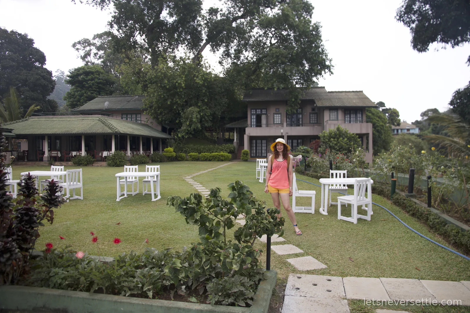 Inga in a sun hat standing among white garden chairs on a guesthouse lawn in Ella, the old bungalow behind