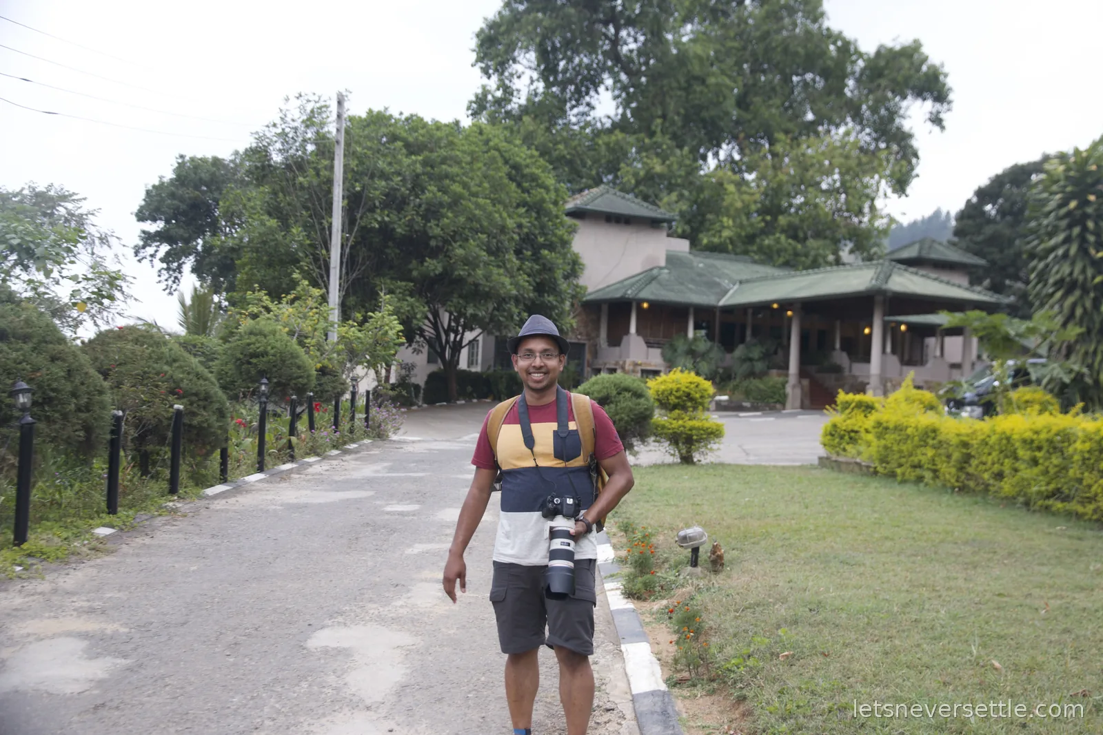 Dileep walking up a driveway carrying his camera, a green roofed guesthouse behind him