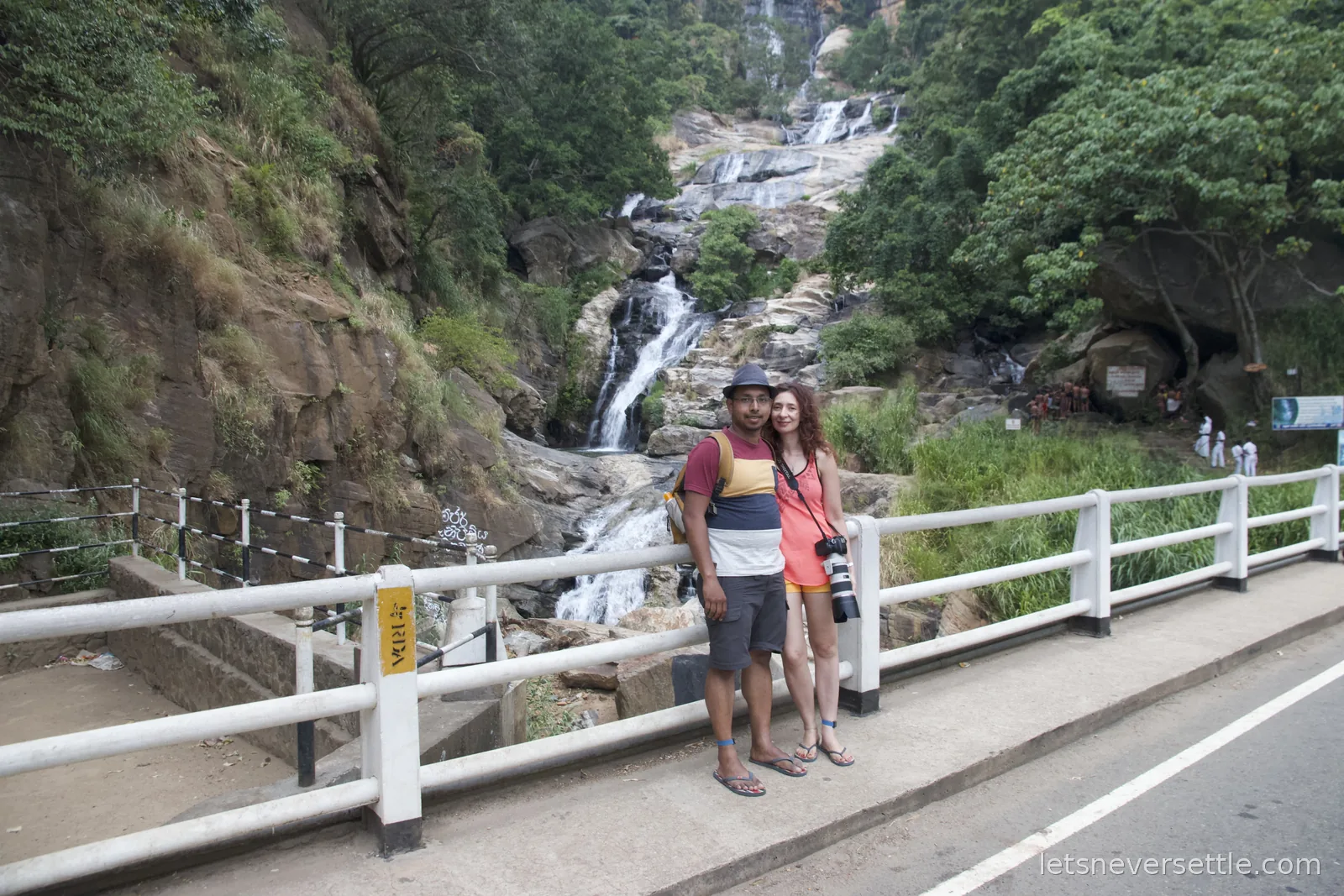 Inga and Dileep standing at a white roadside railing with Ravana Falls tumbling behind them