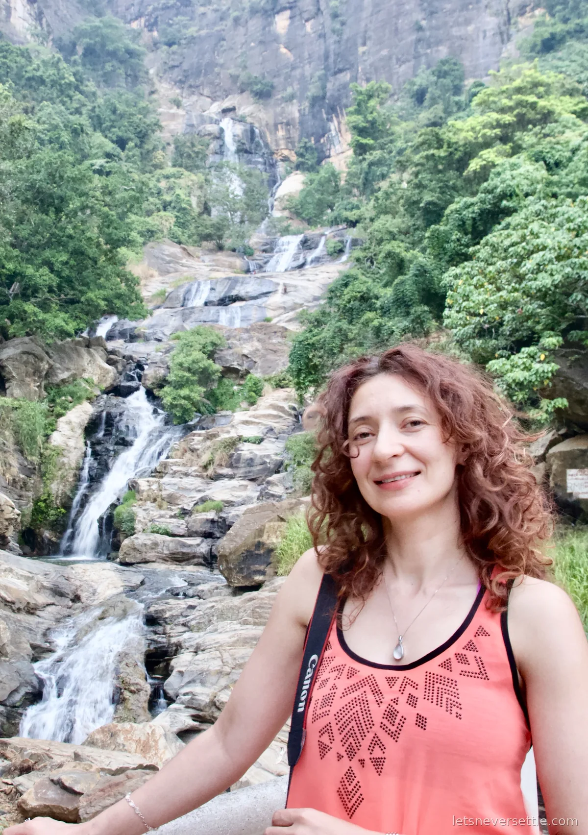 Inga standing in front of the tall cascades of Ravana Falls with a camera strap over one shoulder
