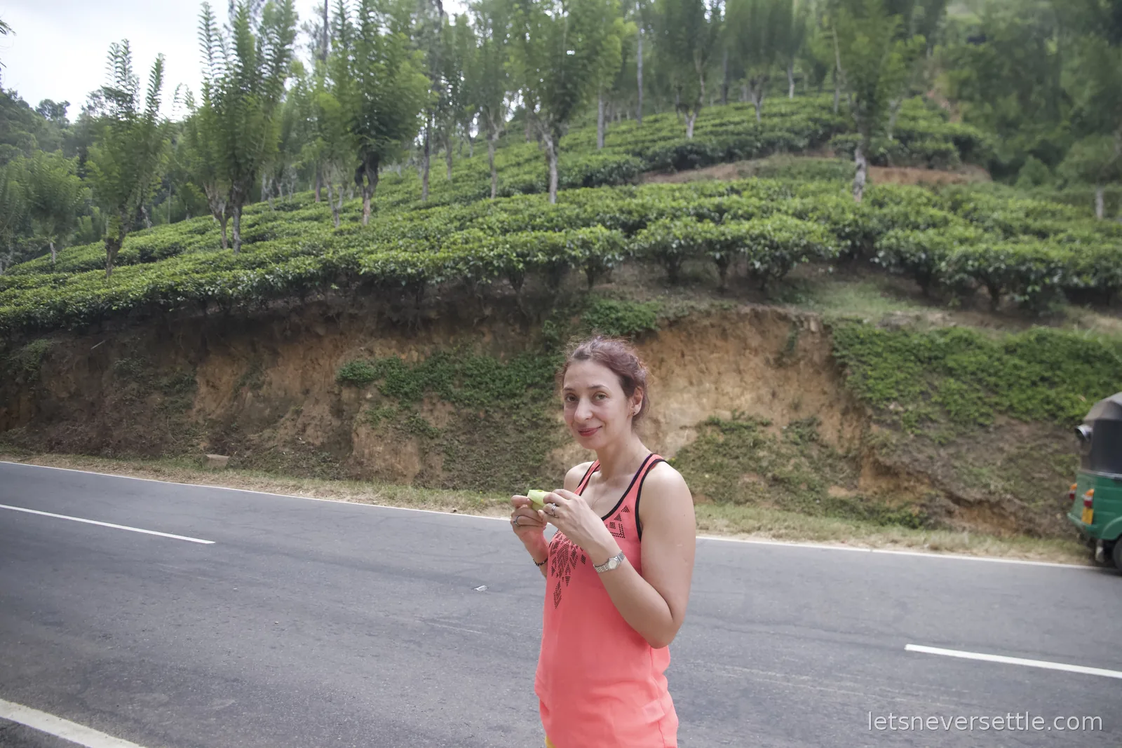 Inga eating a snack beside the road with terraced tea bushes rising on the slope behind her