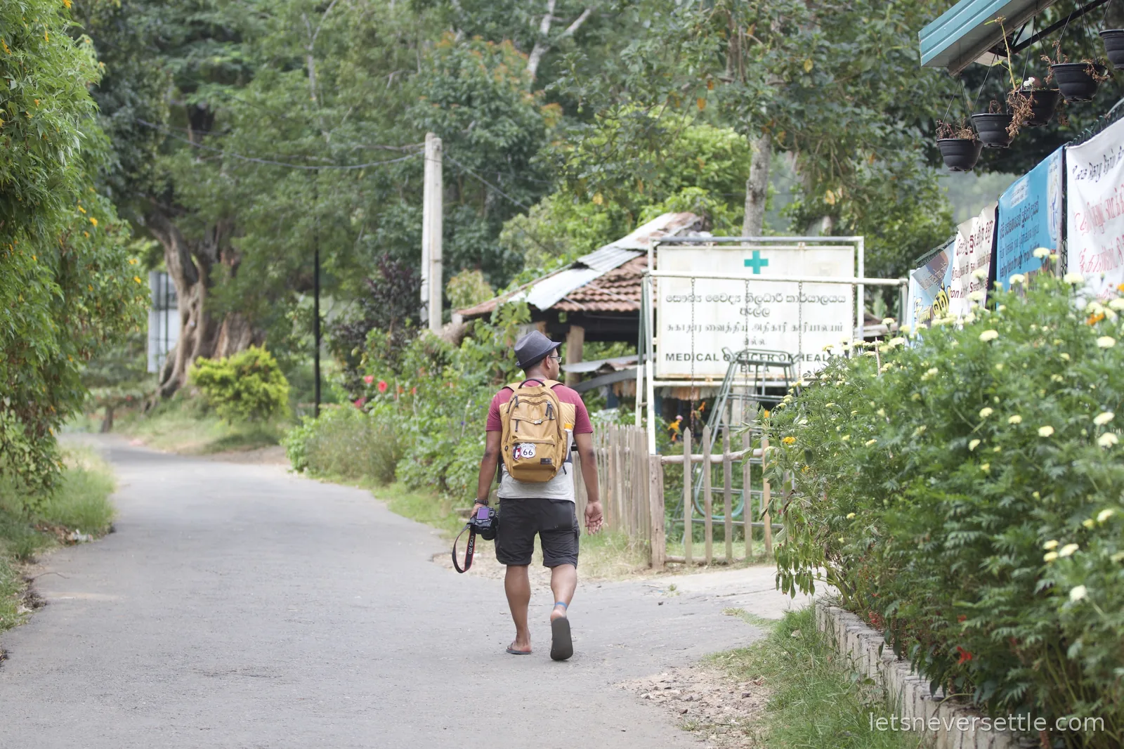 Dileep walking down a quiet lane in Ella past flowering hedges and a green cross sign for the medical officer of health