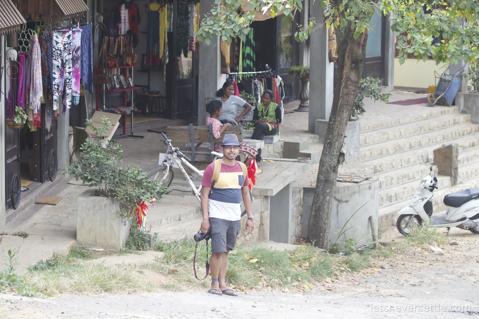 Dileep standing on a dusty verge in Ella village with his camera, clothes shops and women chatting on the steps behind