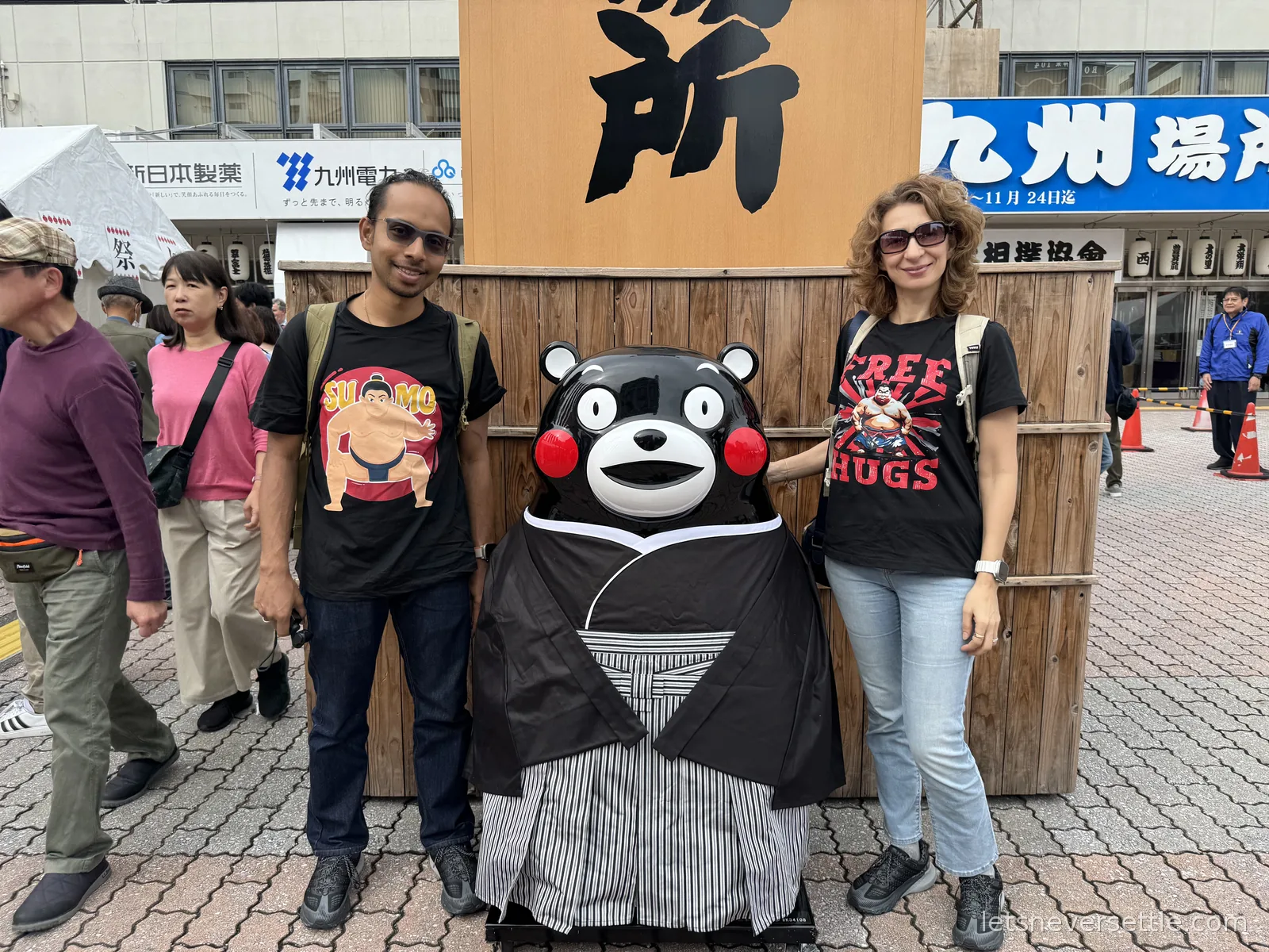 Inga and Dileep in sumo T-shirts posing beside a Kumamon mascot statue dressed in a striped kimono outside the tournament venue