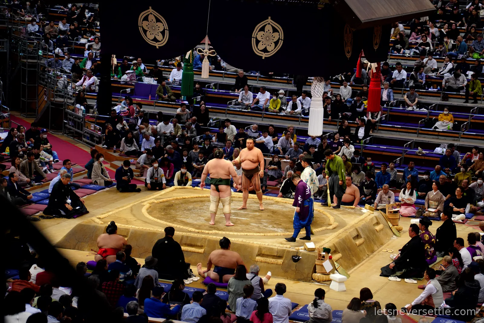 Two sumo wrestlers facing each other in the ring before a bout, watched by a referee in traditional dress and a full crowd