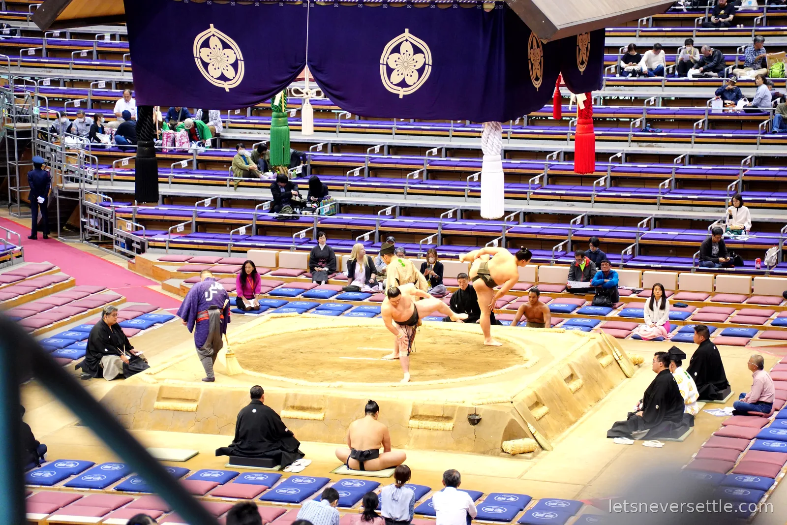 A sumo bout in progress with one wrestler lifting his opponent off balance at the edge of the ring