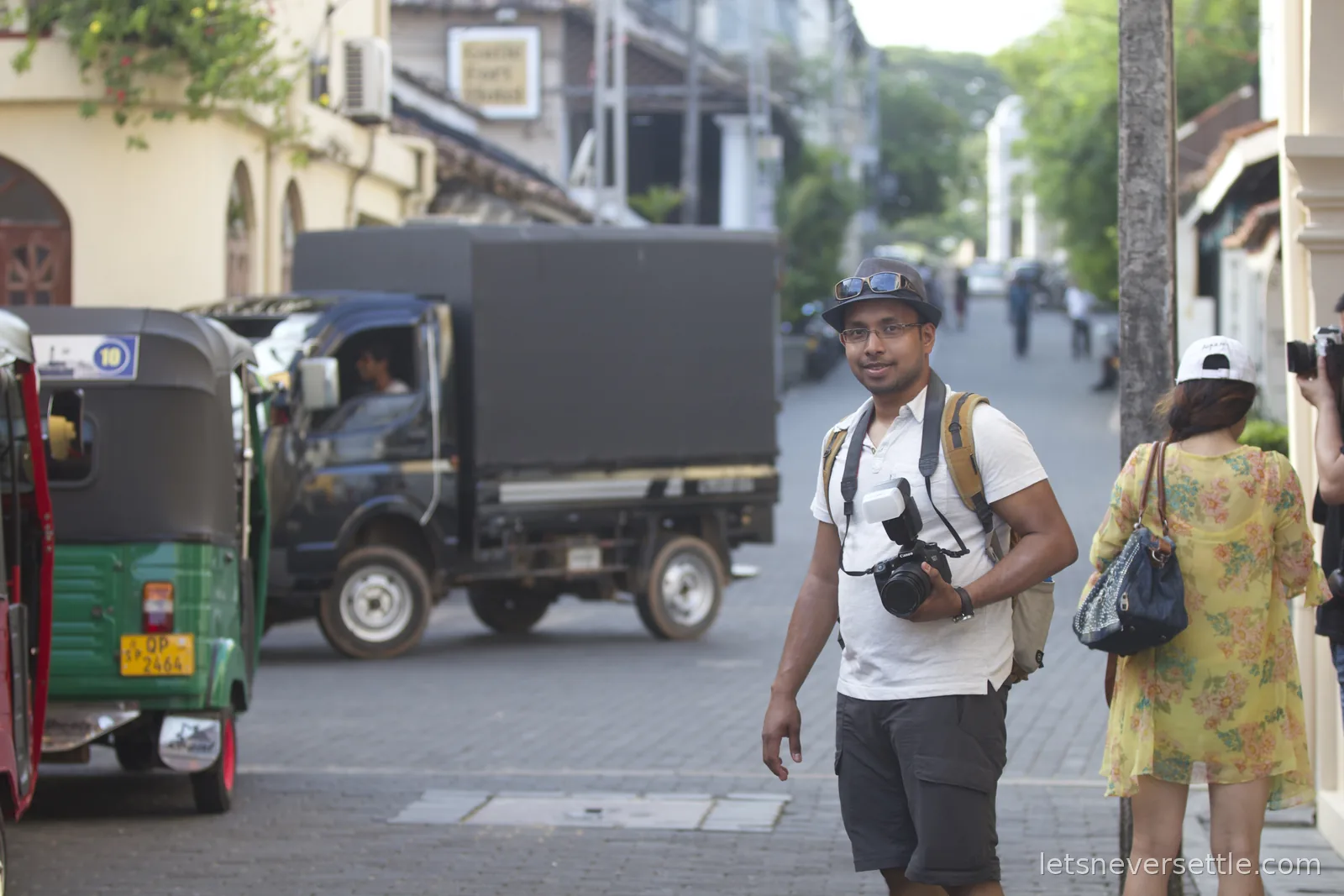 Dileep standing with a camera on a street inside Galle Fort, a green tuk-tuk and a lorry passing behind him