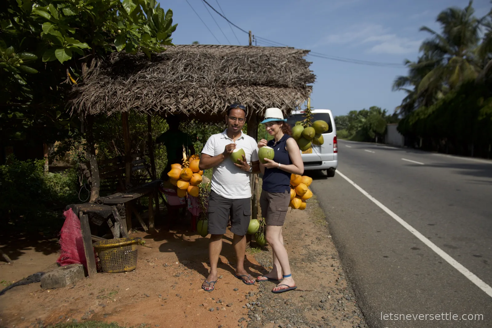Inga and Dileep drinking from king coconuts at a thatched roadside stall hung with bunches of orange coconuts