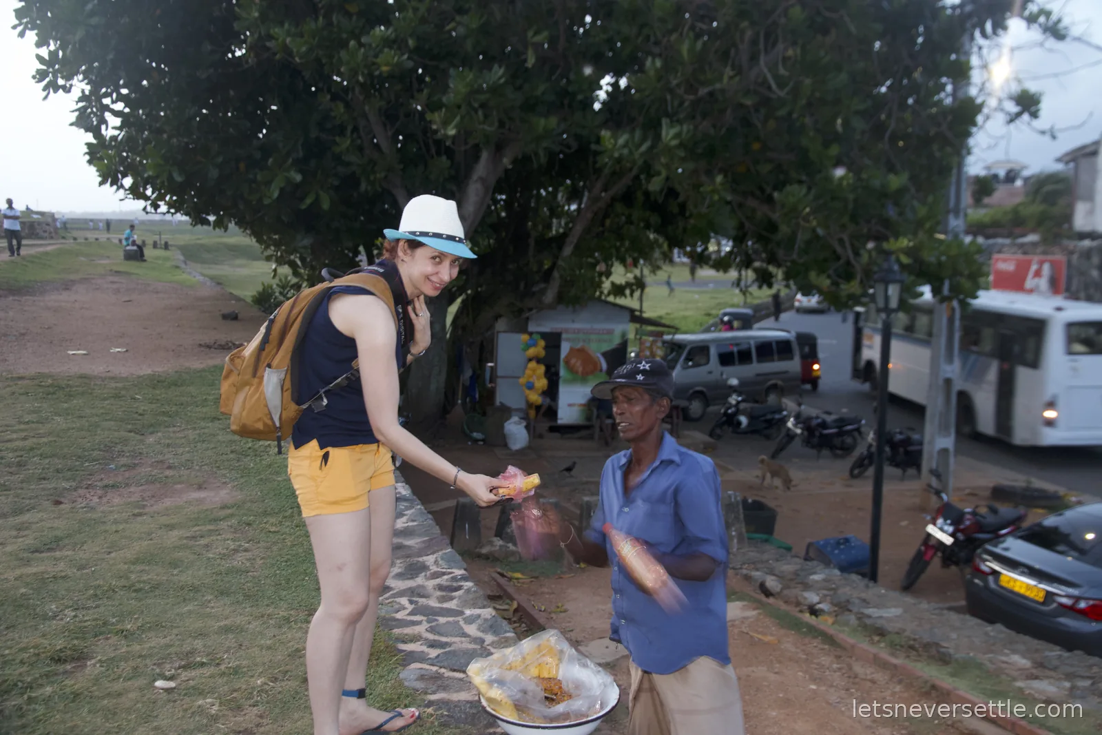 Inga buying a snack from a street vendor with a bowl of fried treats on the fort wall at dusk