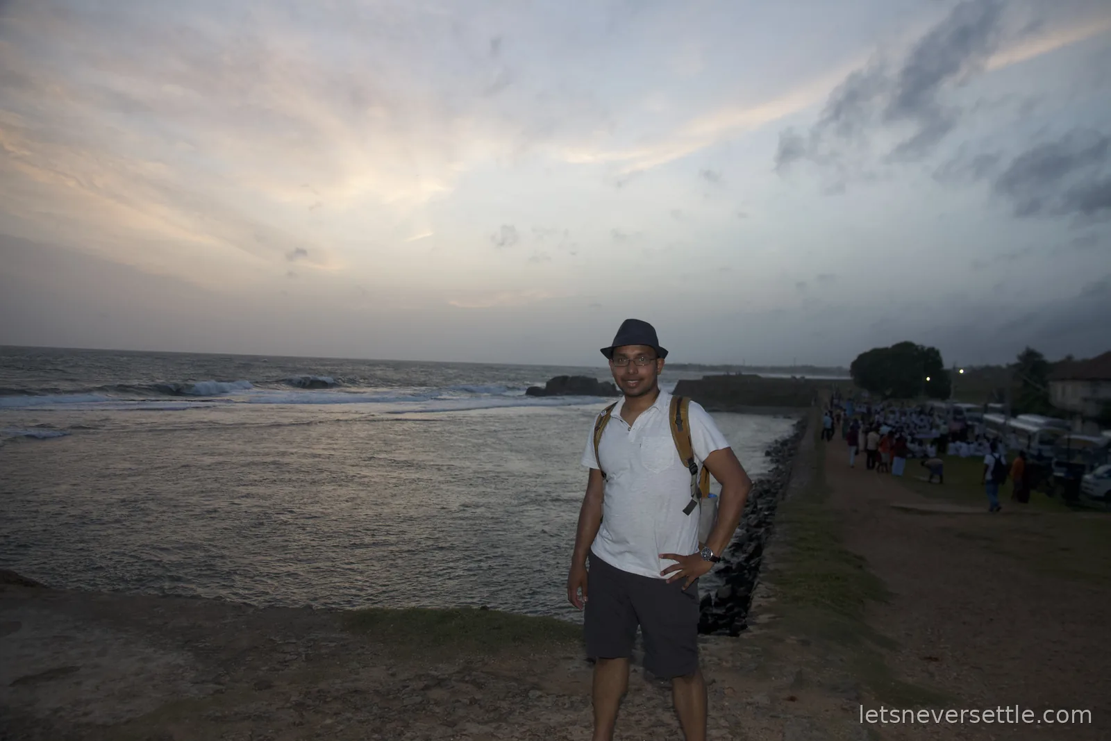 Dileep in a hat standing on the grassy ramparts of Galle Fort at dusk, waves breaking below and crowds along the wall