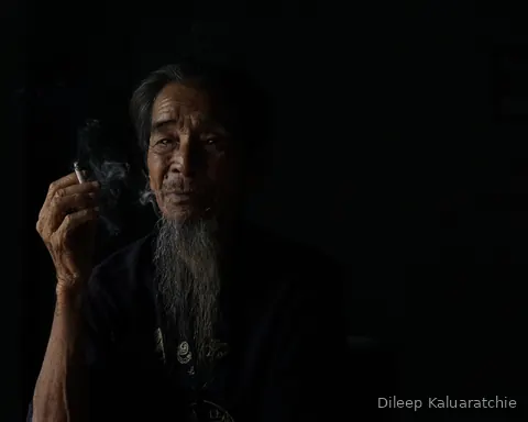 An elderly man with a long grey beard drawing on a cigarette, the smoke caught in the light