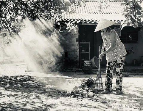 Black and white: a woman in a conical hat raking burning leaves in a smoky yard outside a tiled house