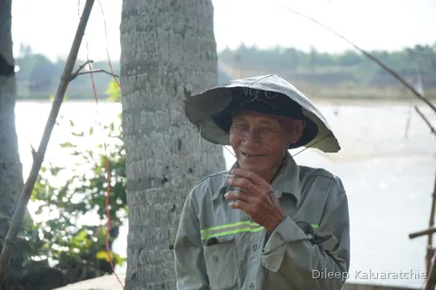 A smiling older man in a conical hat and work shirt, resting against a palm trunk by the water