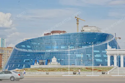 A curved blue glass building in Astana with construction cranes behind it