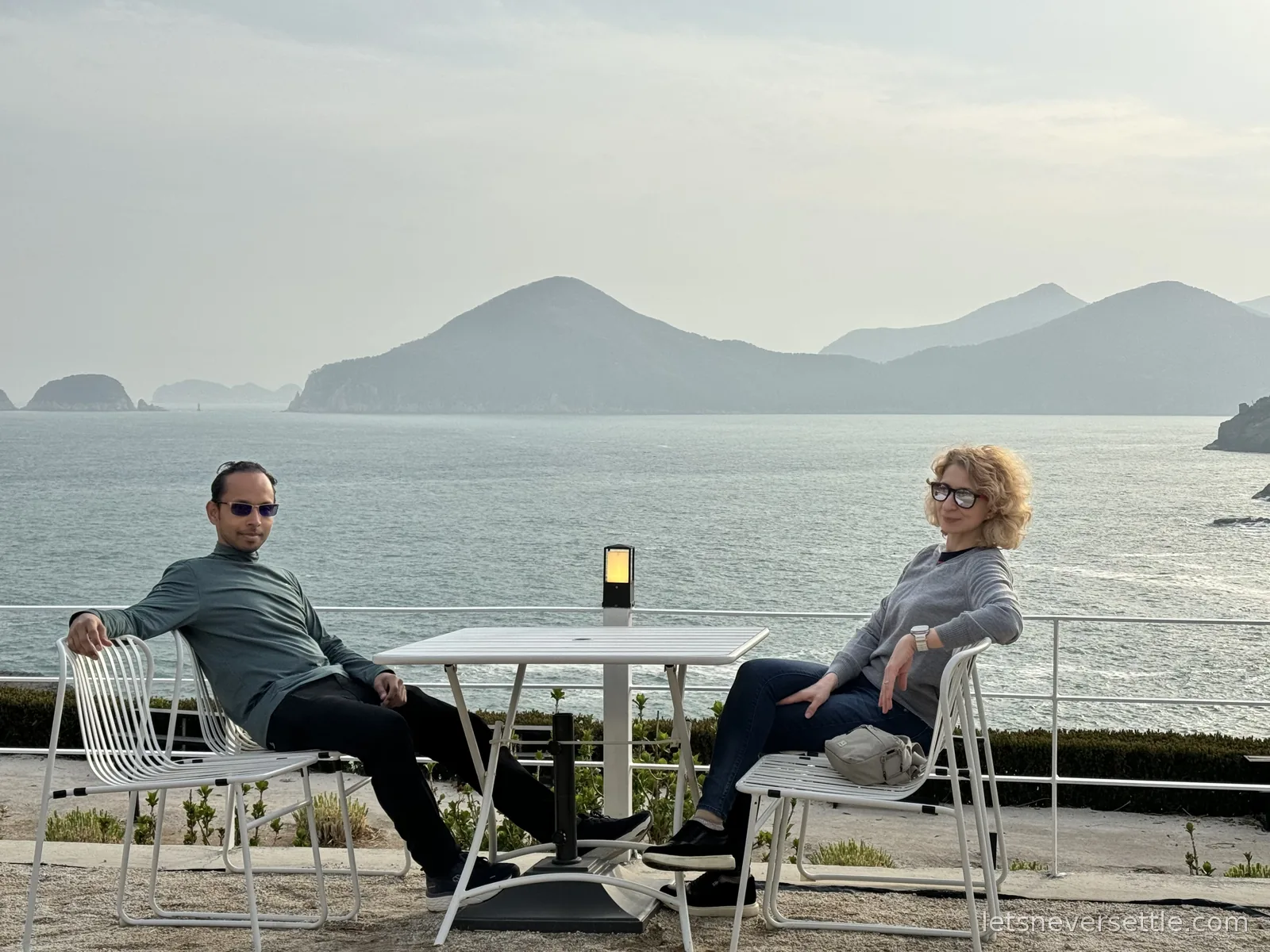 Inga and Dileep lounging on white chairs at a terrace table above the sea on Geoje, layers of hazy islands behind