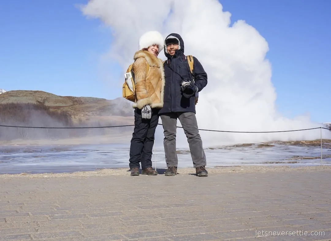 Inga and Dileep posing on the path at Geysir while a huge white column of steam erupts behind them
