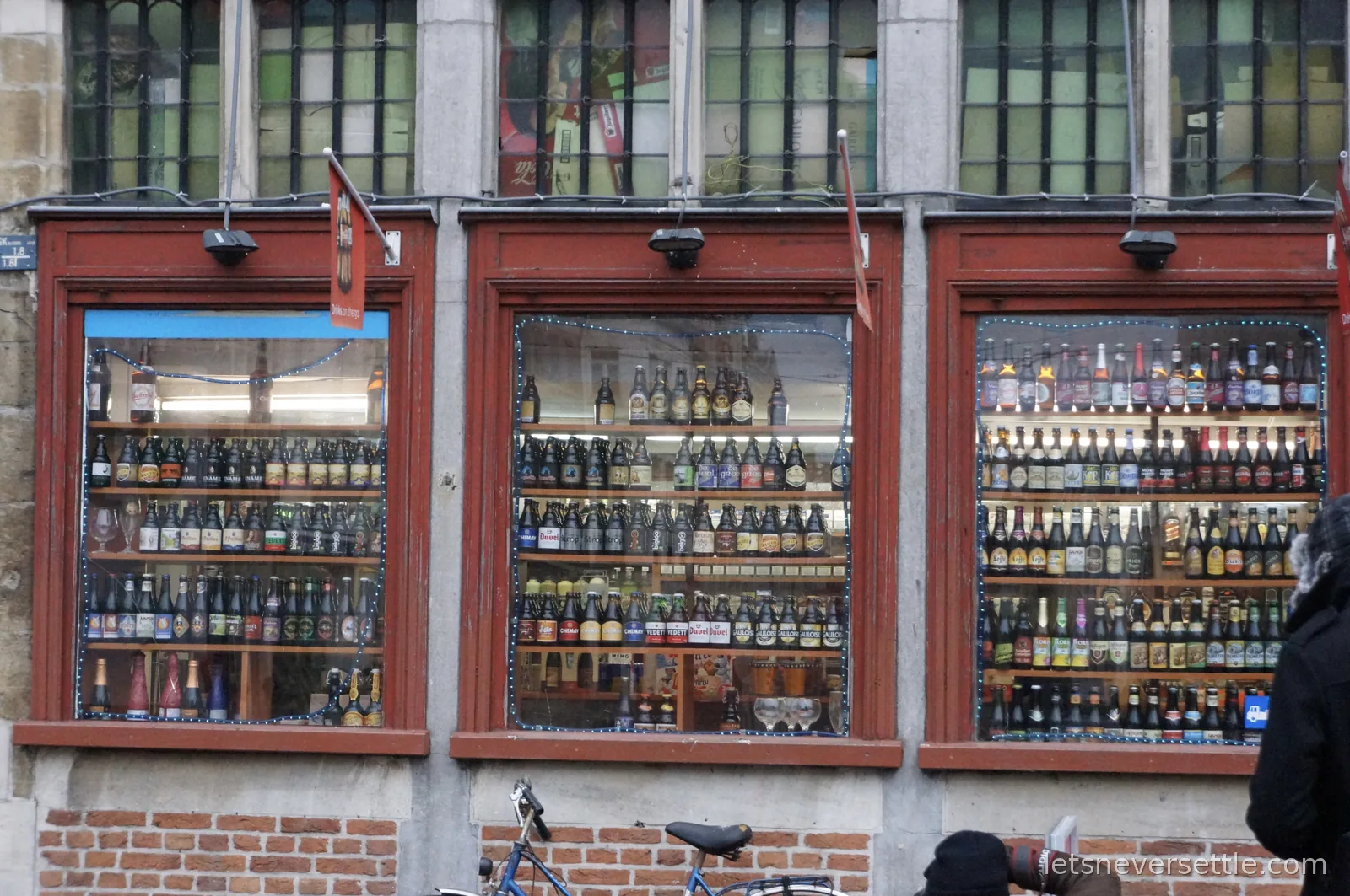Three shop windows in Ghent filled with shelves of Belgian beer bottles, with bicycles parked outside