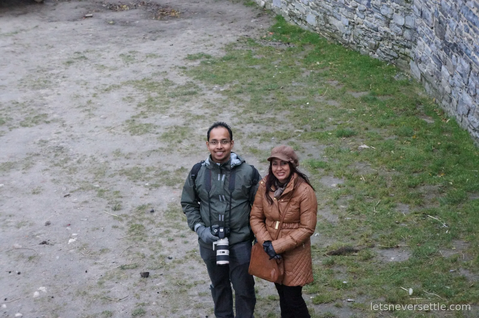 A man with a camera and a woman in a tan winter coat smiling up from the grounds of Gravensteen castle, photographed from the wall above